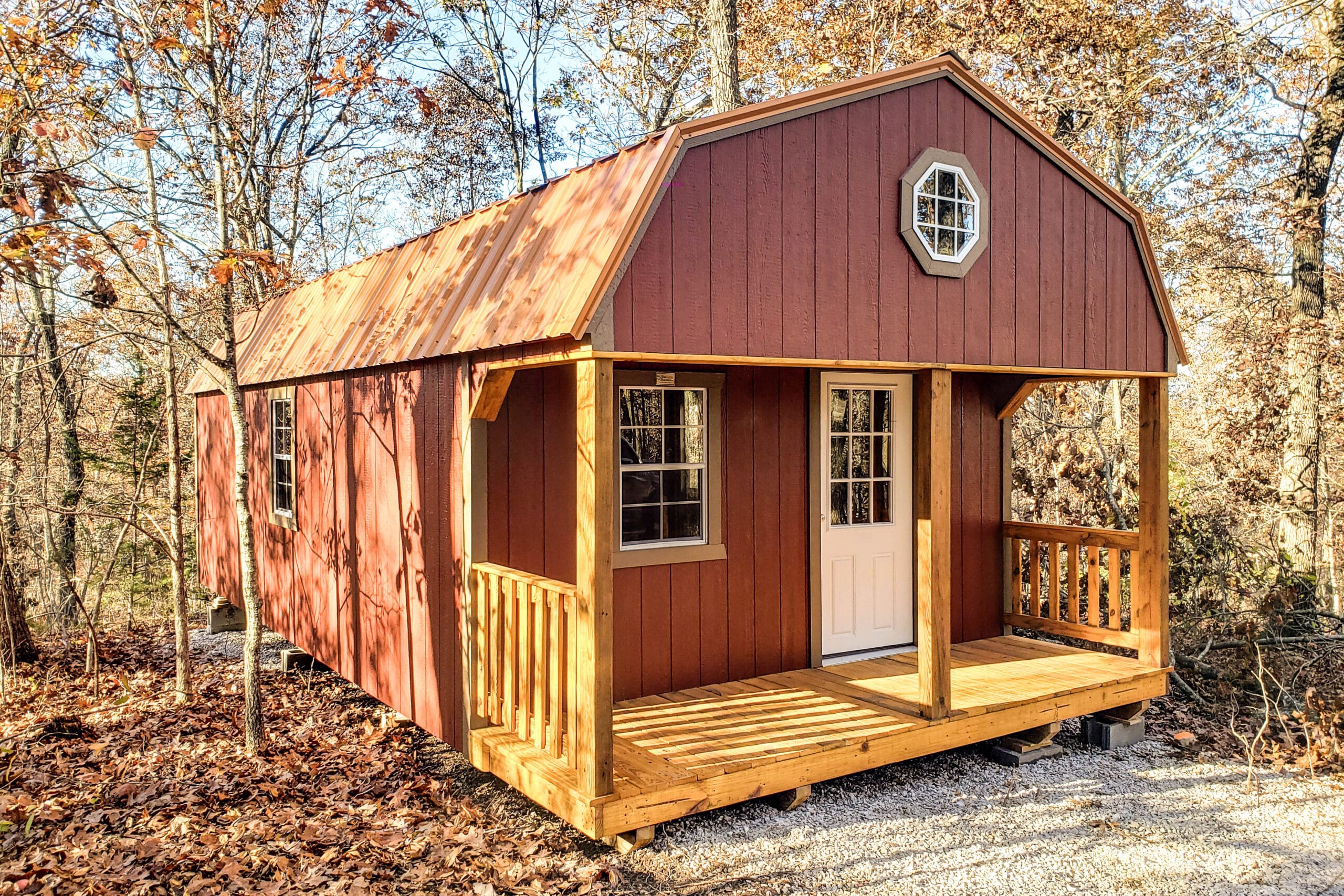 One of our red lofted cabins sheds for sale in Sullivan MO- Red siding whith fenced porch four visible white windows and windowed door beneath metal gray roof