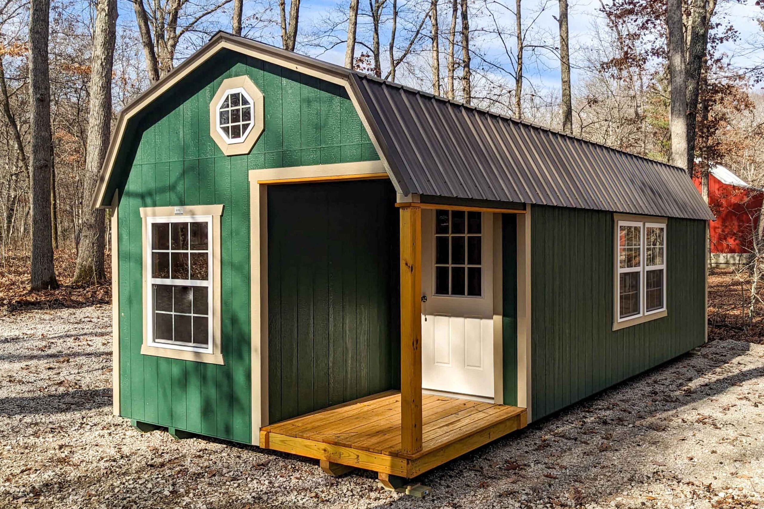 One of our red lofted cabins sheds for sale in Sullivan MO- Red siding whith fenced porch four visible white windows and windowed door beneath metal gray roof