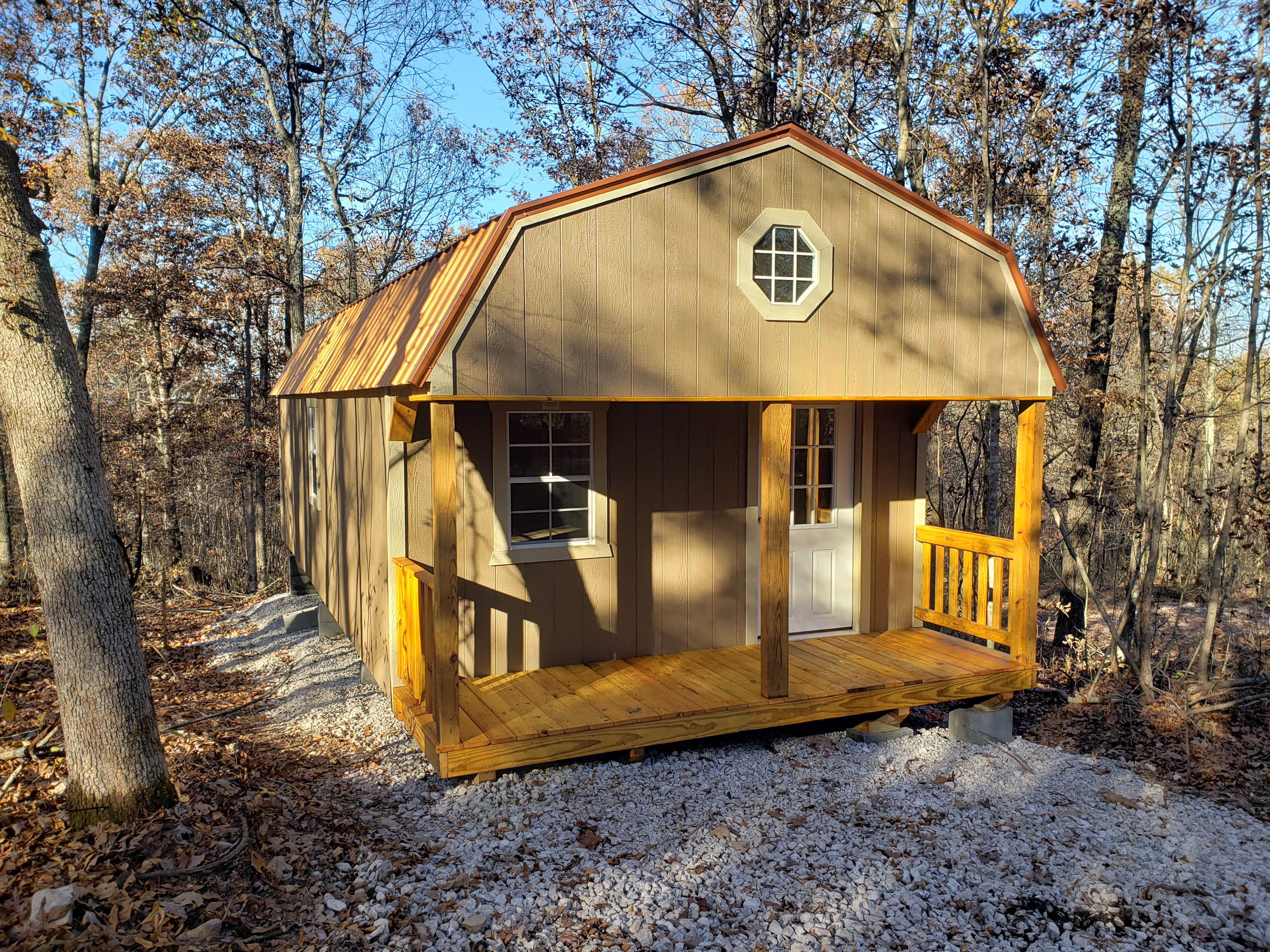 One of our red lofted cabins sheds for sale in Sullivan MO- Red siding whith fenced porch four visible white windows and windowed door beneath metal gray roof