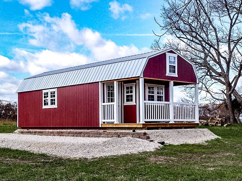 One of our red lofted cabins sheds for sale in Sullivan MO- Red siding whith fenced porch four visible white windows and windowed door beneath metal gray roof