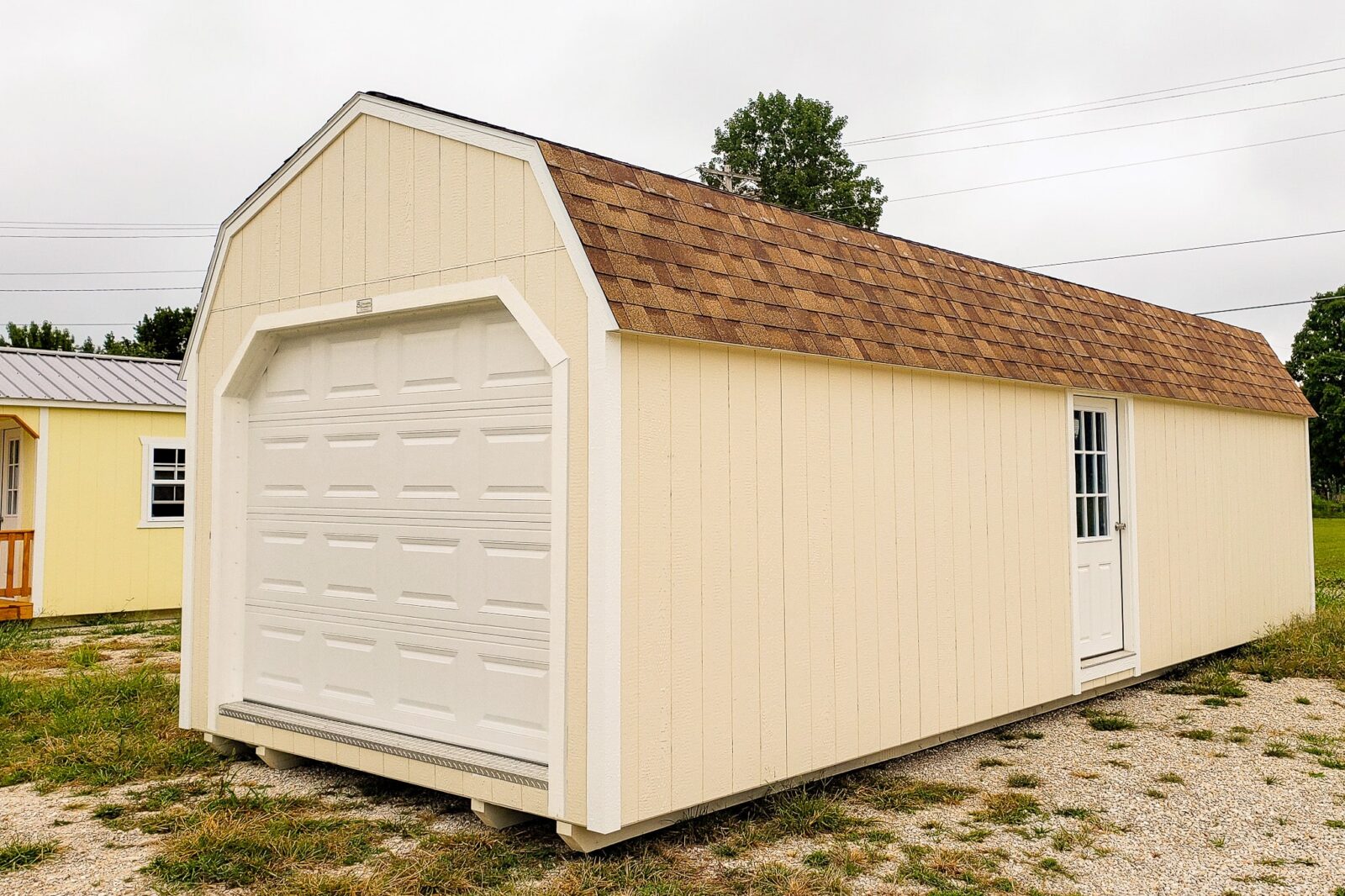 One of our light beige lofted garage sheds in Sullivan MO - Light beige siding and white overhead and single side door under brown shingled roof on field of patch grass and nearby yellow shed on cloudy day