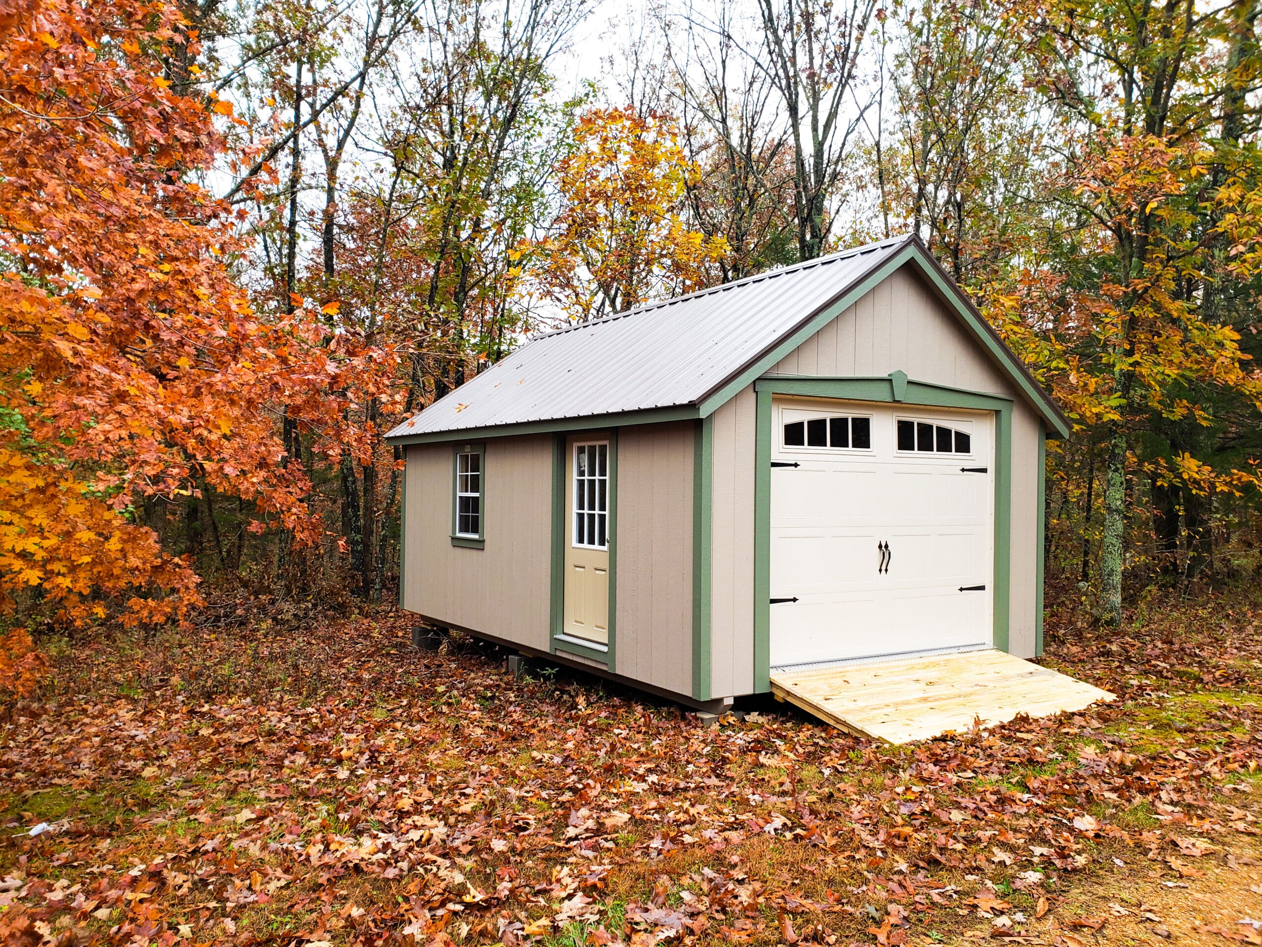 One of our popular tan and green-trimmed garages for sale near Washington MO - Prefab garage shed sits among bed of autumn leaves near shedding trees